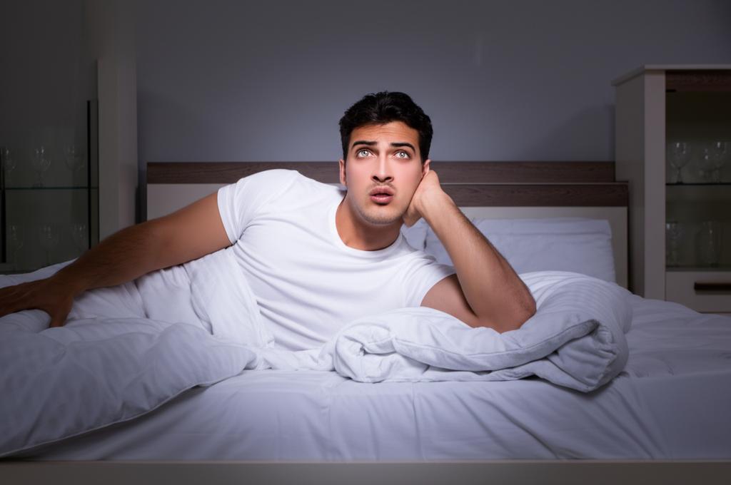 A stressed man lying in bed at night, unable to sleep, with a worried expression on his face.